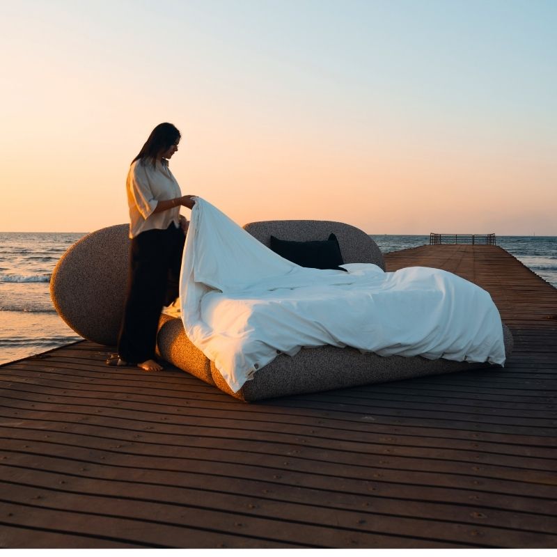 Minimalist organic shaped bed with a stone-like texture and curved headboard on a wooden pier at sunset. Mahogni