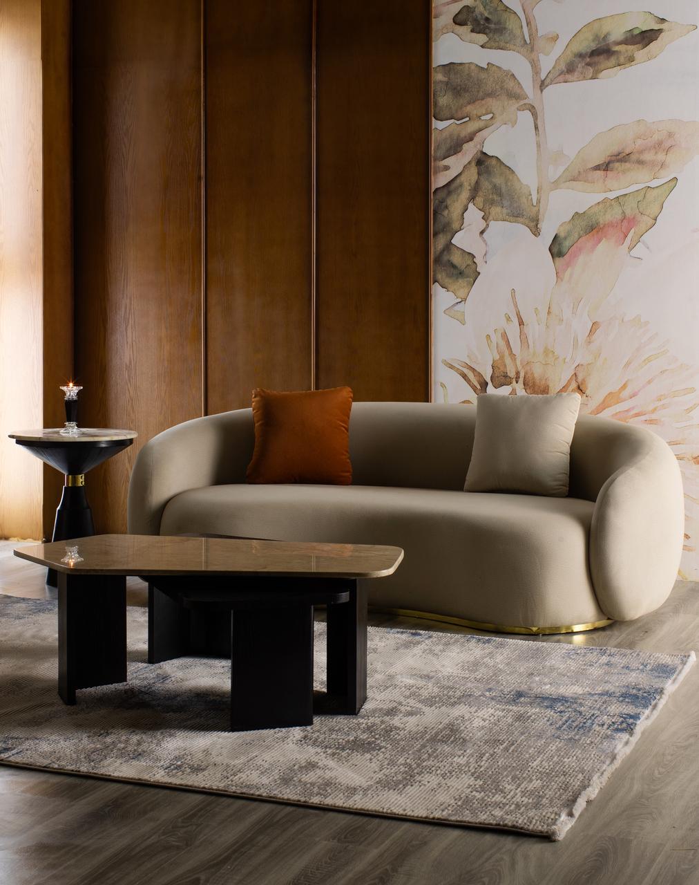 Modern curved sofa in sand beige velvet with a gold base, featuring a burnt orange accent pillow and a beige cushion, styled on a contemporary rug in an elegant room setting.