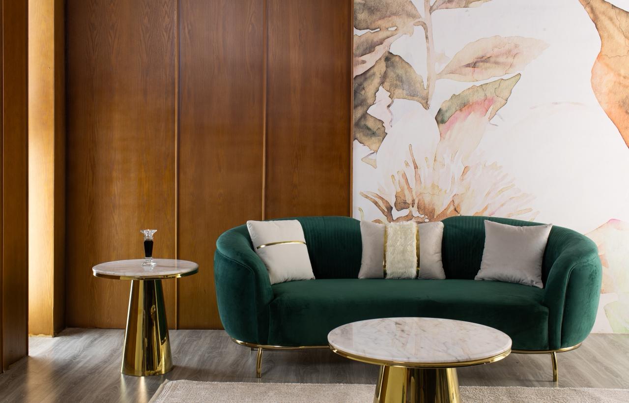 Luxurious curved sofa upholstered in emerald green velvet with a polished gold metal base, styled with decorative cushions and marble coffee tables in an elegant living room.