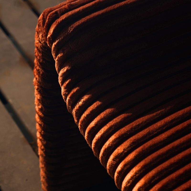 Rust orange ribbed velvet armchair with a sculptural design on a wooden deck by the sea.