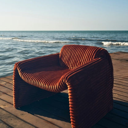 Rust orange ribbed velvet armchair with a sculptural design on a wooden deck by the sea. Mahogni