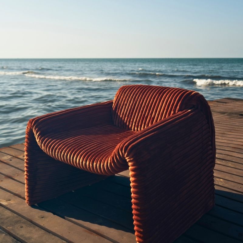 Rust orange ribbed velvet armchair with a sculptural design on a wooden deck by the sea. Mahogni