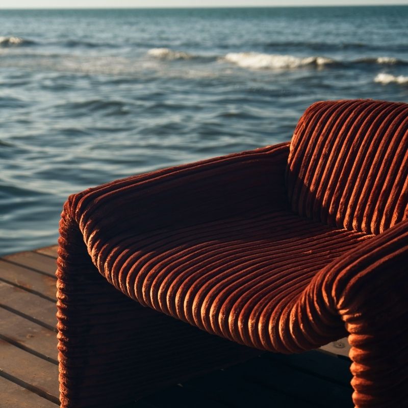 Rust orange ribbed velvet armchair with a sculptural design on a wooden deck by the sea. Mahogni