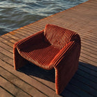 Rust orange ribbed velvet armchair with a sculptural design on a wooden deck by the sea. Mahogni