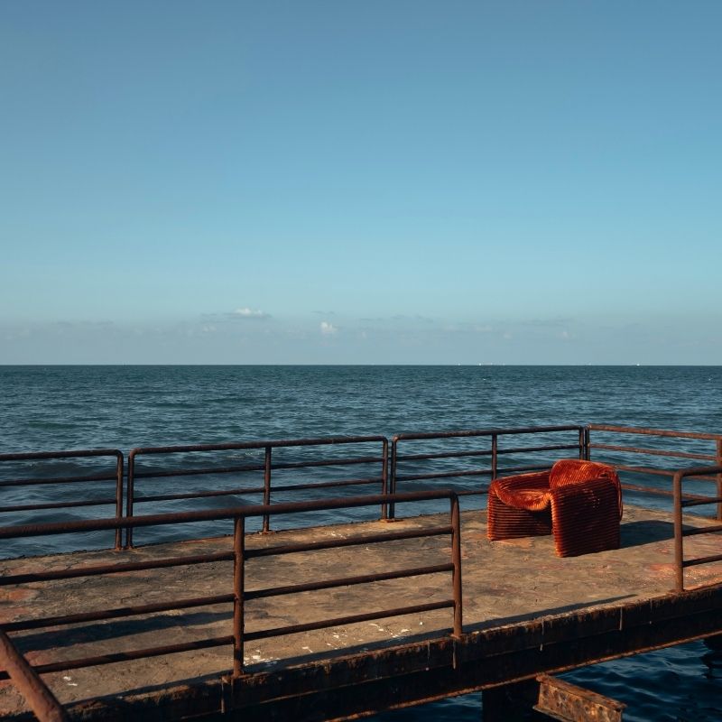 Rust orange ribbed velvet armchair with a sculptural design on a wooden deck by the sea. Mahogni