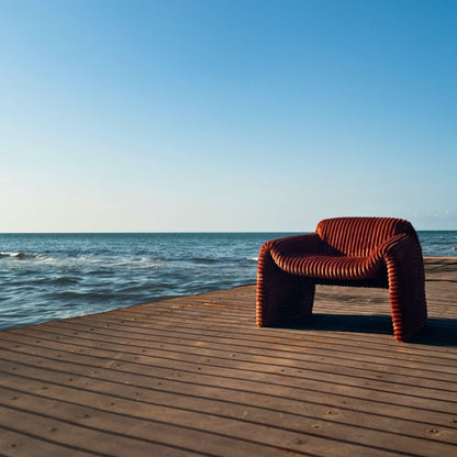 Rust orange ribbed velvet armchair with a sculptural design on a wooden deck by the sea. Mahogni