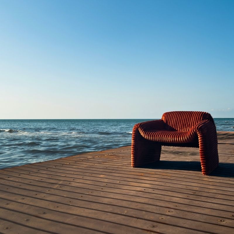 Rust orange ribbed velvet armchair with a sculptural design on a wooden deck by the sea. Mahogni