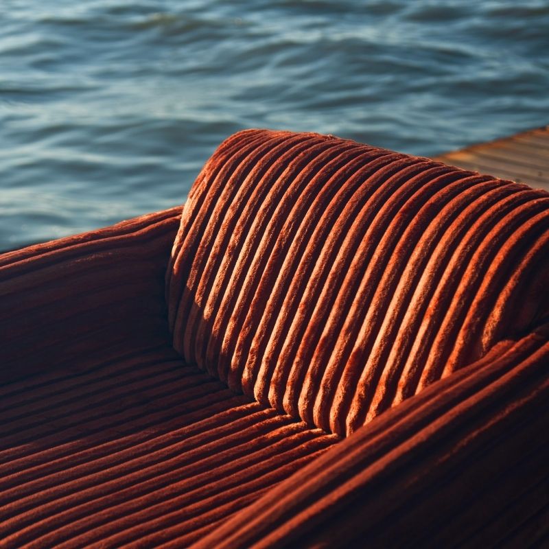 Rust orange ribbed velvet armchair with a sculptural design on a wooden deck by the sea. Mahogni