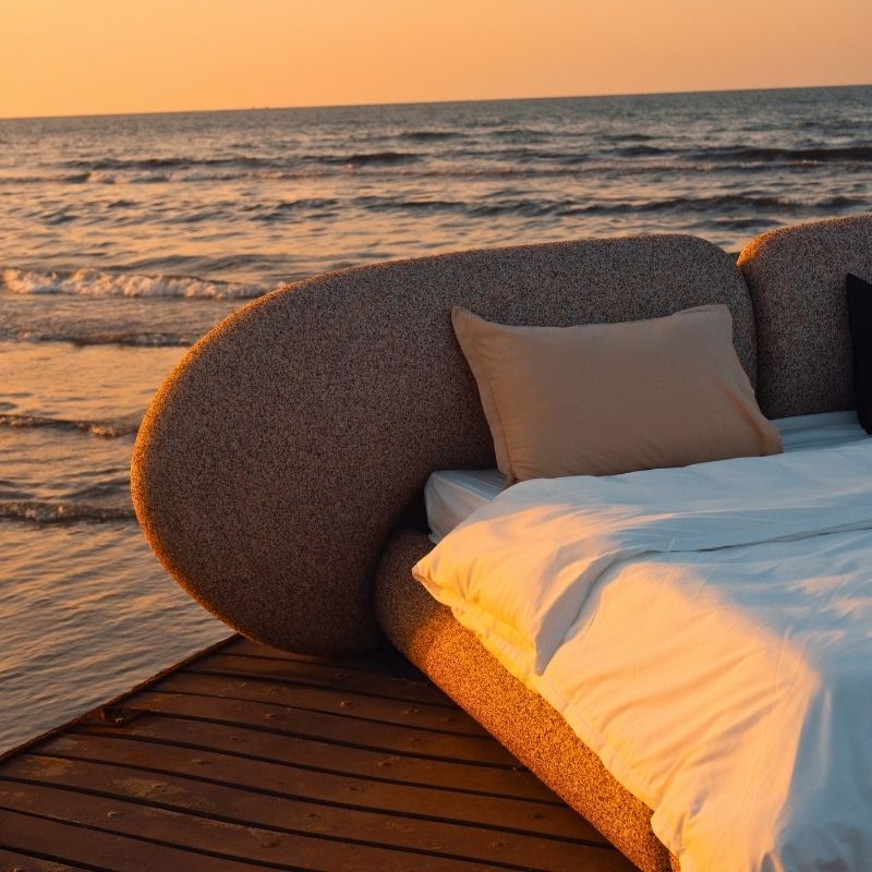 Minimalist organic shaped bed with a stone-like texture and curved headboard on a wooden pier at sunset. Mahogni 