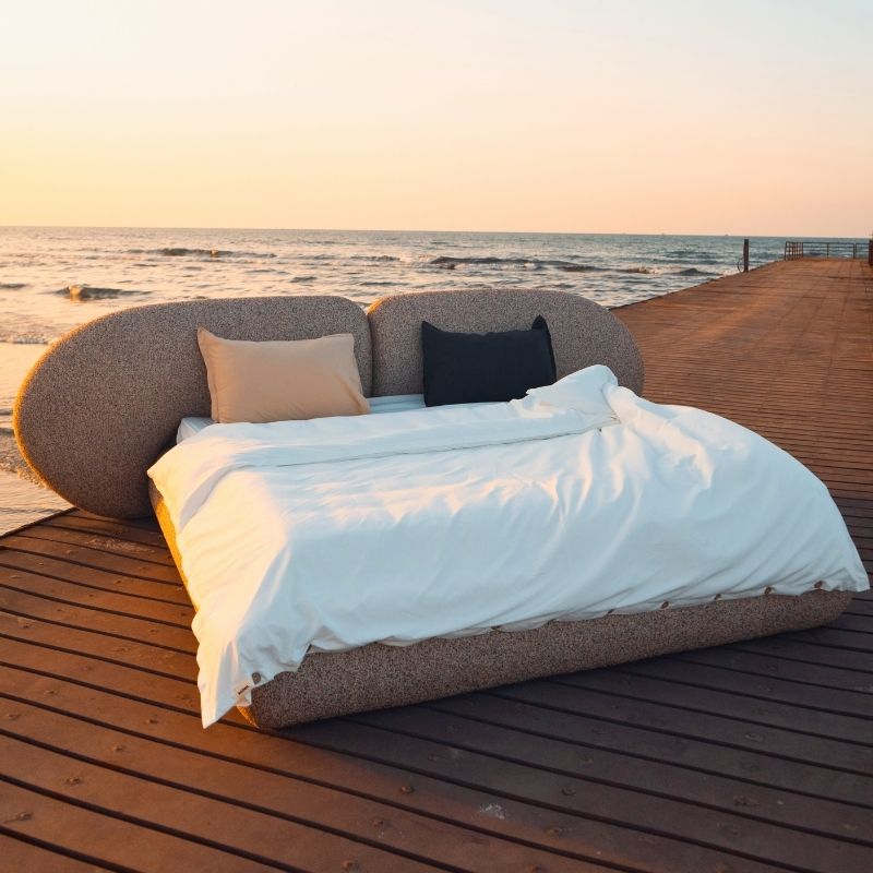 Minimalist organic shaped bed with a stone-like texture and curved headboard on a wooden pier at sunset. Mahogni 