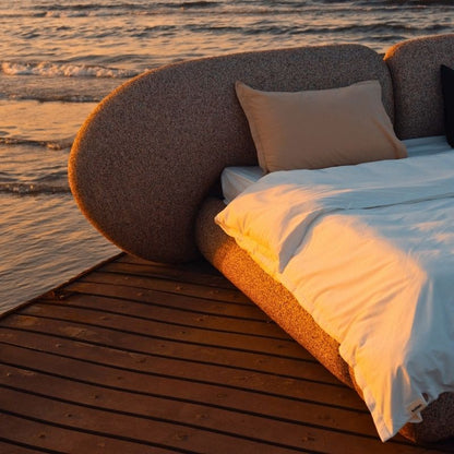 Minimalist organic shaped bed with a stone-like texture and curved headboard on a wooden pier at sunset. Mahogni 
