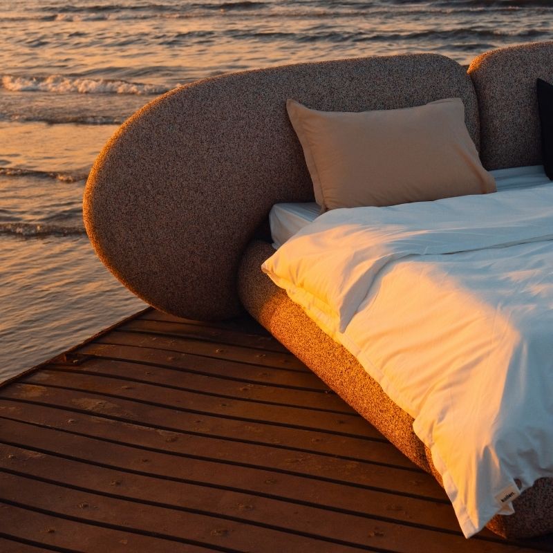 Minimalist organic shaped bed with a stone-like texture and curved headboard on a wooden pier at sunset. Mahogni 