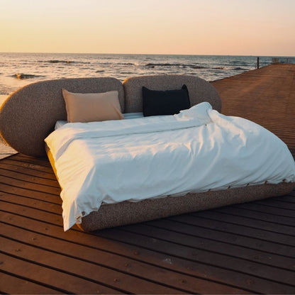 Minimalist organic shaped bed with a stone-like texture and curved headboard on a wooden pier at sunset. Mahogni 