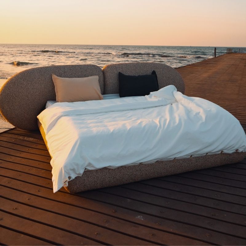 Minimalist organic shaped bed with a stone-like texture and curved headboard on a wooden pier at sunset. Mahogni 