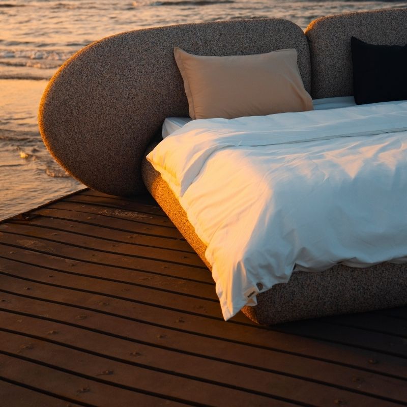 Minimalist organic shaped bed with a stone-like texture and curved headboard on a wooden pier at sunset. Mahogni 