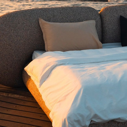 Minimalist organic shaped bed with a stone-like texture and curved headboard on a wooden pier at sunset. Mahogni 