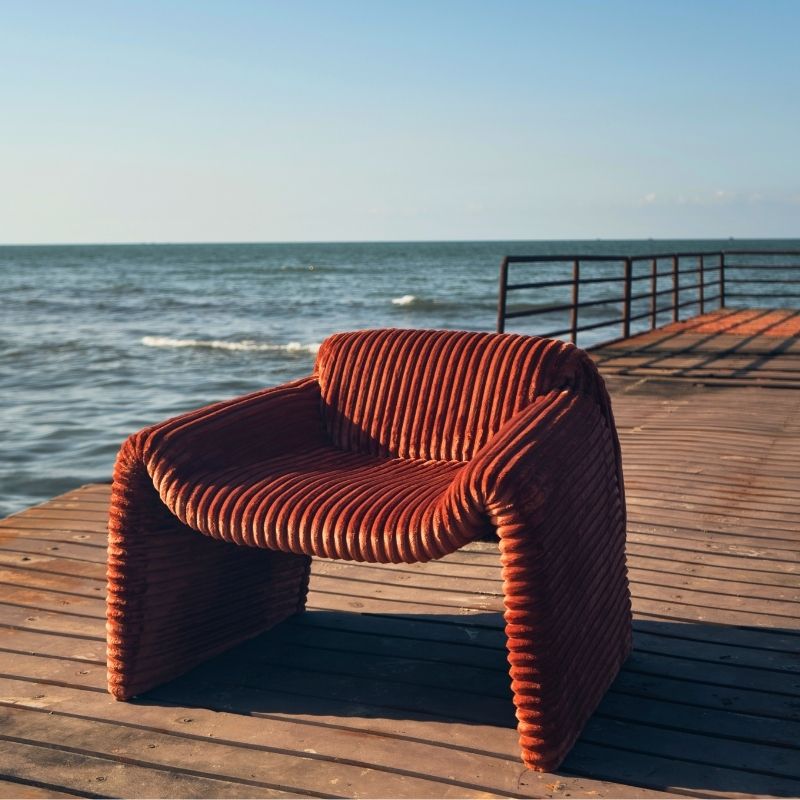 Rust orange ribbed velvet armchair with a sculptural design on a wooden deck by the sea. Mahogni
