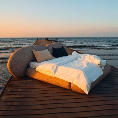 Minimalist organic shaped bed with a stone-like texture and curved headboard on a wooden pier at sunset. Mahogni 