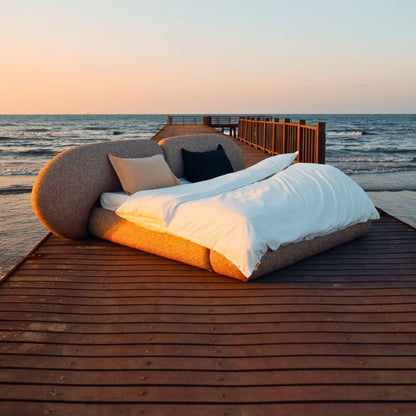 Minimalist organic shaped bed with a stone-like texture and curved headboard on a wooden pier at sunset. Mahogni 