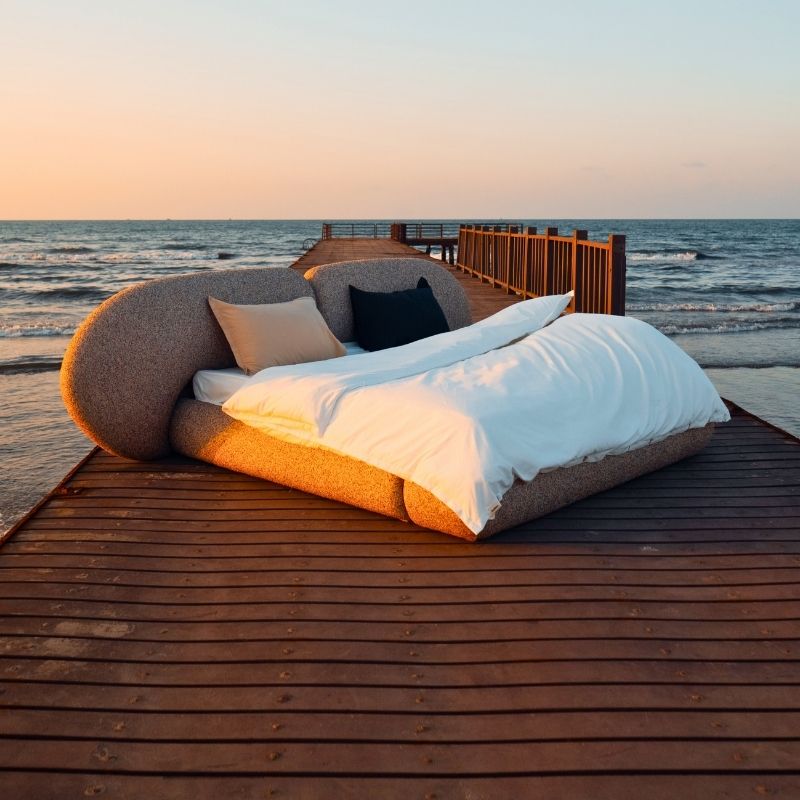 Minimalist organic shaped bed with a stone-like texture and curved headboard on a wooden pier at sunset. Mahogni 