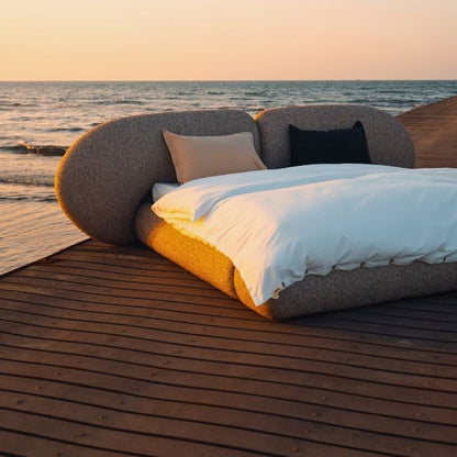 Minimalist organic shaped bed with a stone-like texture and curved headboard on a wooden pier at sunset. Mahogni 