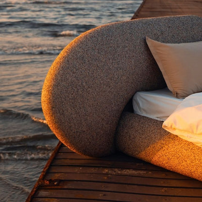 Minimalist organic shaped bed with a stone-like texture and curved headboard on a wooden pier at sunset. Mahogni 