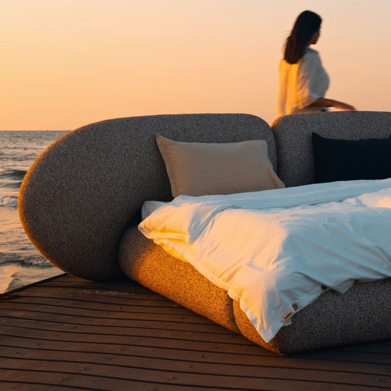 Minimalist organic shaped bed with a stone-like texture and curved headboard on a wooden pier at sunset. Mahogni 