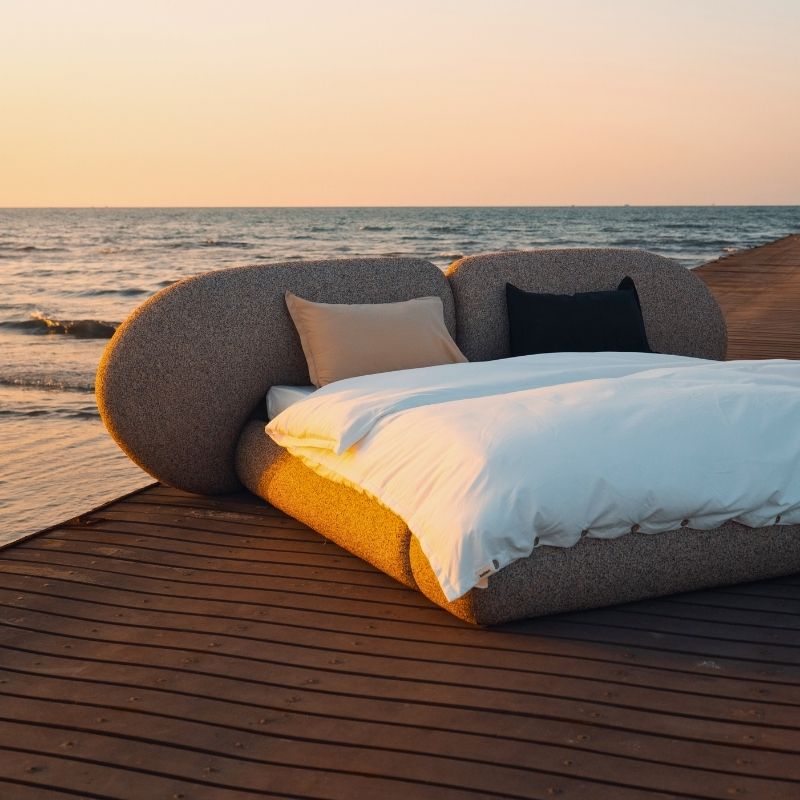 Minimalist organic shaped bed with a stone-like texture and curved headboard on a wooden pier at sunset. Mahogni 