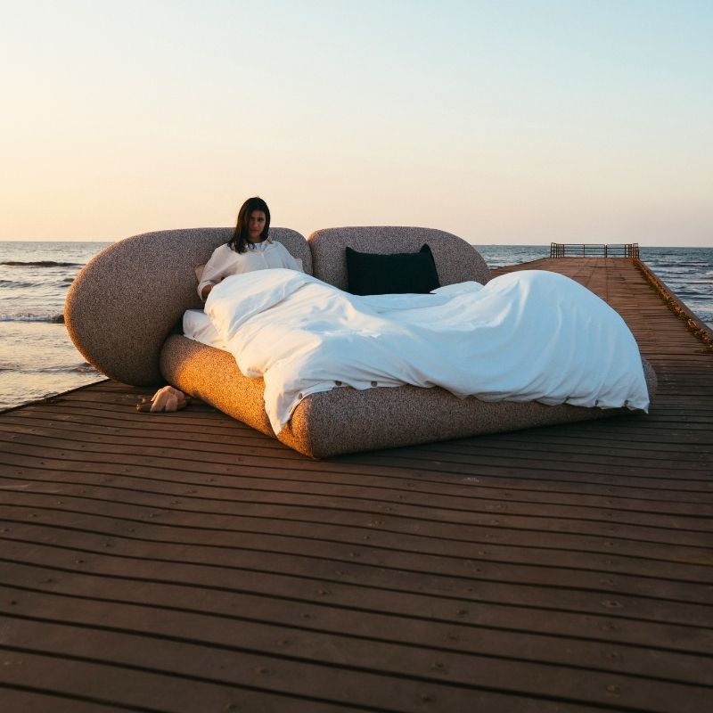 Minimalist organic shaped bed with a stone-like texture and curved headboard on a wooden pier at sunset. Mahogni