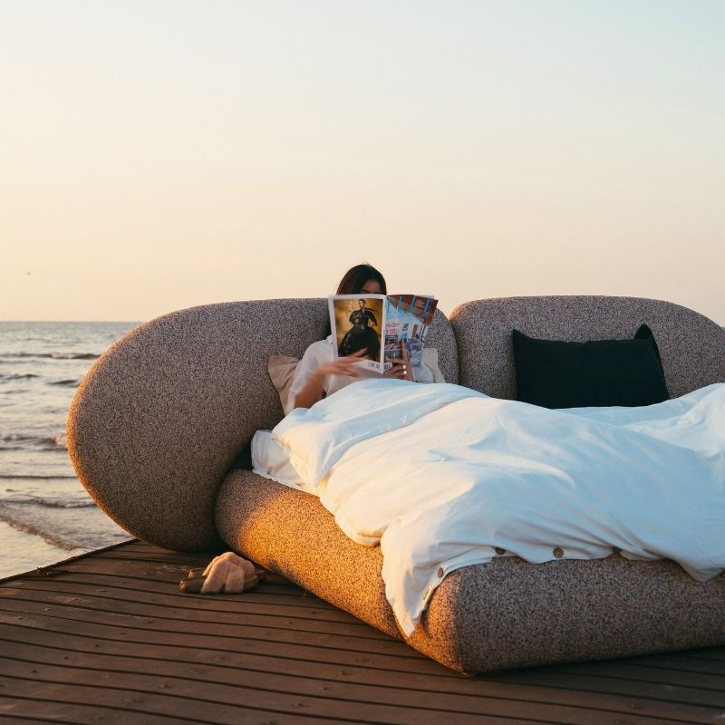 Minimalist organic shaped bed with a stone-like texture and curved headboard on a wooden pier at sunset. Mahogni 