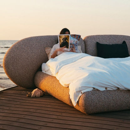 Minimalist organic shaped bed with a stone-like texture and curved headboard on a wooden pier at sunset. Mahogni 