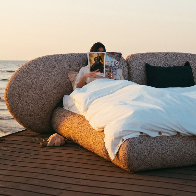 Minimalist organic shaped bed with a stone-like texture and curved headboard on a wooden pier at sunset. Mahogni 