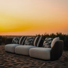 Complete modular living room sofa set in pebble grey with charcoal geometric pillows on a sandy backdrop at sunset. Mahogni 