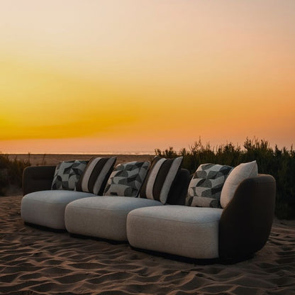 Complete modular living room sofa set in pebble grey with charcoal geometric pillows on a sandy backdrop at sunset. Mahogni 
