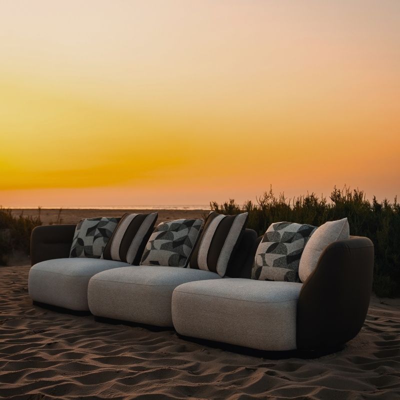 Complete modular living room sofa set in pebble grey with charcoal geometric pillows on a sandy backdrop at sunset. Mahogni 