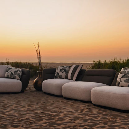 Complete modular living room sofa set in pebble grey with charcoal geometric pillows on a sandy backdrop at sunset. Mahogni 