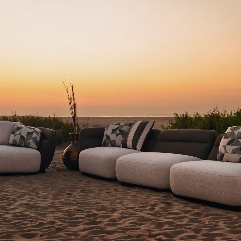 Complete modular living room sofa set in pebble grey with charcoal geometric pillows on a sandy backdrop at sunset. Mahogni 
