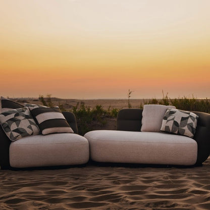 Complete modular living room sofa set in pebble grey with charcoal geometric pillows on a sandy backdrop at sunset. Mahogni 
