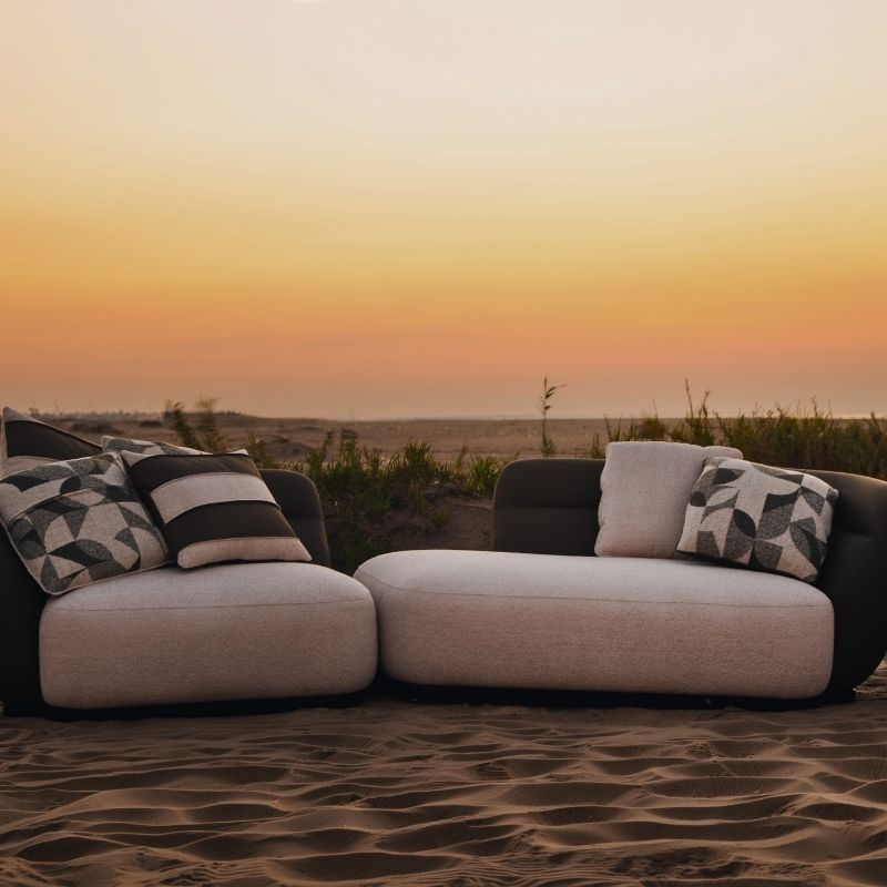 Complete modular living room sofa set in pebble grey with charcoal geometric pillows on a sandy backdrop at sunset. Mahogni 