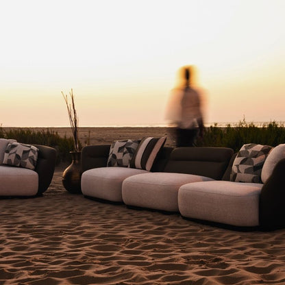 Complete modular living room sofa set in pebble grey with charcoal geometric pillows on a sandy backdrop at sunset. Mahogni 