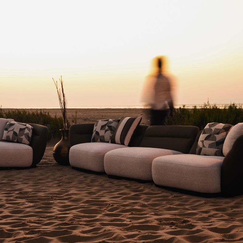 Complete modular living room sofa set in pebble grey with charcoal geometric pillows on a sandy backdrop at sunset. Mahogni 